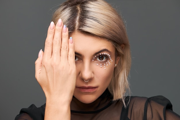 rätselhafte trendige junge europäische Frau mit blond gefärbten Haaren und Kristallen im Gesicht als Teil des Make-ups, ein Auge mit Handfläche bedeckend, polierte Nägel zeigend. Kunst und Kosmetik