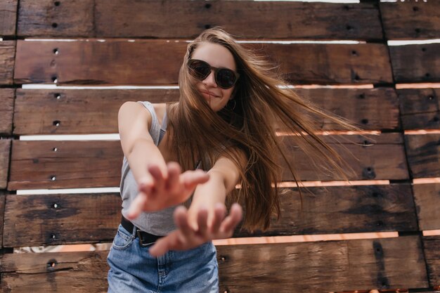 Raffinierte europäische Frau in der stilvollen Sonnenbrille, die auf Holzwand tanzt. Porträt des anmutigen Mädchens mit der langen Frisur, die Glück im warmen Sommertag ausdrückt.