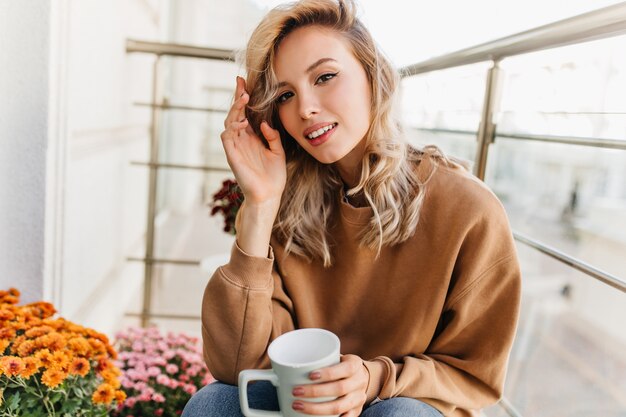 Raffinierte junge Frau, die Tee am Balkon trinkt. Hübsches blondes Mädchen, das Kaffee am Morgen genießt.