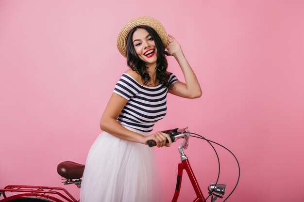 Raffinierte lateinamerikanische Frau im gestreiften T-Shirt, das emotional aufwirft. Wunderschönes brünettes Mädchen im Hut, das mit Fahrrad steht.