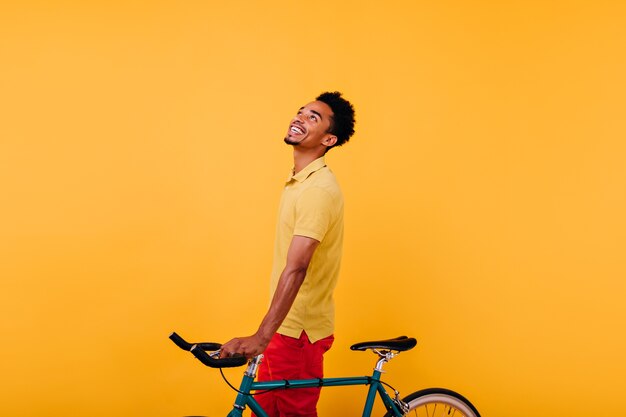 Raffinierter afrikanischer Mann, der mit Fahrrad nach oben schaut. Innenfoto des sorglosen schwarzen Kerls, der Spaß hat.