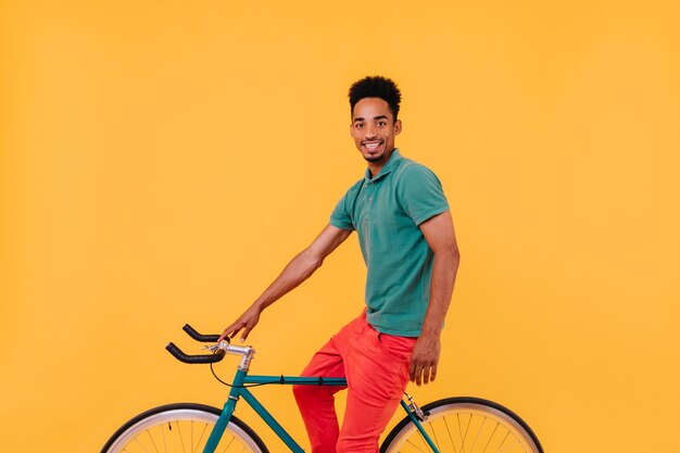 Raffinierter männlicher Radfahrer, der mit Lächeln aufwirft. Interessierter brünetter Kerl, der auf Fahrrad nahe gelber Wand sitzt.