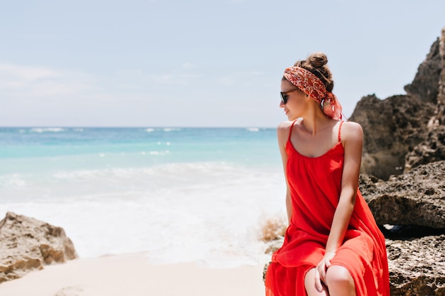 Raffiniertes kaukasisches weibliches Modell, das auf Stein sitzt und Meerblick genießt. Romantische weiße junge Frau, die Ozean durch Sonnenbrille betrachtet.