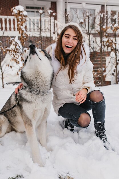 Raffiniertes weibliches Modell in warmer Kleidung, die während der Winterferien mit Husky-Hund herumalbert. Außenporträt der atemberaubenden jungen Dame spielt mit Haustier im Dezembermorgen.