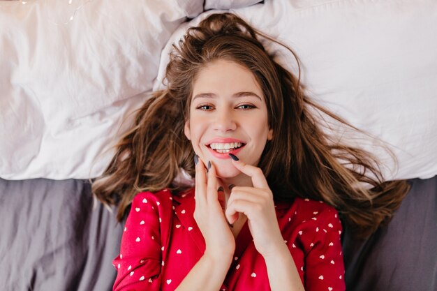 Raffiniertes weißes Mädchen, das positive Gefühle ausdrückt, während es im Bett liegt. Innenfoto der fröhlichen braunhaarigen Frau lächelnd
