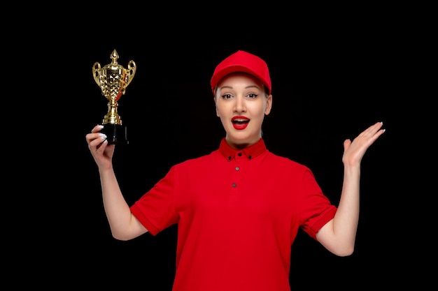 Red Shirt Day aufgeregtes Siegermädchen, das eine Trophäe in einer roten Kappe mit Hemd und hellem Lippenstift hält