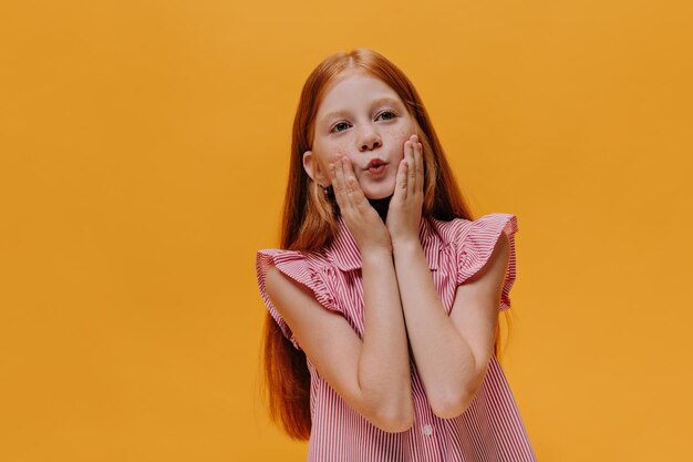 Redhead hübsches Mädchen in rot gestreiftem Hemd berührt ihre Wangen und pfeift Charmantes Kind posiert auf orangefarbenem, isoliertem Hintergrund