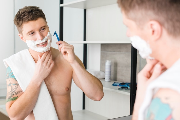 Reflexion des lächelnden jungen Mannes, der im Spiegel rasiert mit Rasiermesser schaut