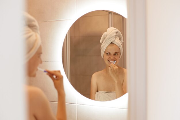 Reflexion im Spiegel einer gewinnenden jungen erwachsenen Frau mit weißem Handtuch auf ihrem Haar, die im Badezimmer posiert und sich die Zähne putzt, Hygieneverfahren nach dem Duschen.