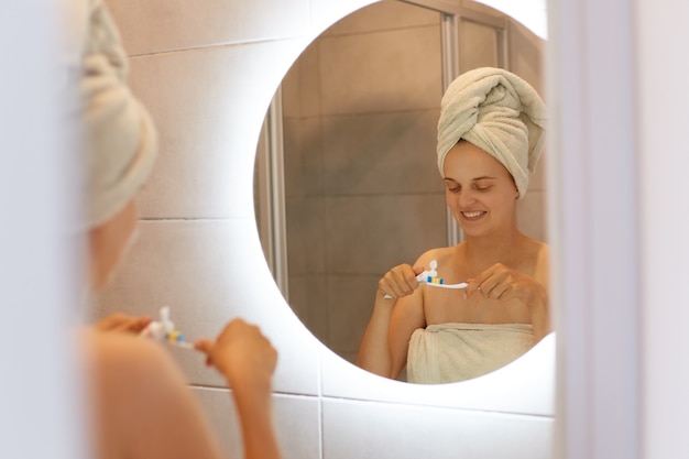 Kostenloses Foto reflexion im spiegel einer glücklichen, positiven jungen frau, die nach dem duschen in ein handtuch gewickelt ist, zahnpasta auf die zahnbürste aufträgt, zahnhygiene, morgenroutine.