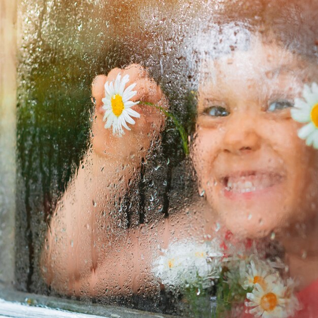 Regentropfen auf dem Glas eines Dorffensters, Kamille blüht Augen in Kinderhänden schauen auf den Regen.