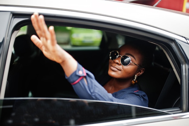 Reiche afrikanische Geschäftsfrau mit Sonnenbrille sitzt am Geländewagen und zeigt Hand aus dem Fenster