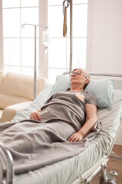 Reife einsame Frau, die in einem mit einer Decke bedeckten Pflegeheim auf dem Bett liegt.