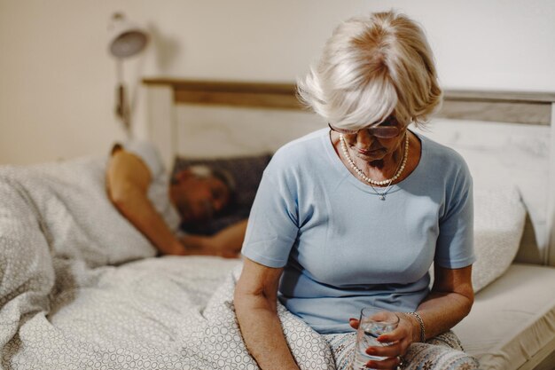 Reife Frau, die auf dem Bett sitzt und ein Glas Wasser hält Ihr Mann schläft im Hintergrund