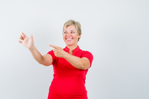 Reife Frau, die auf ihre erhabene Handfläche im roten T-Shirt zeigt und froh aussieht.