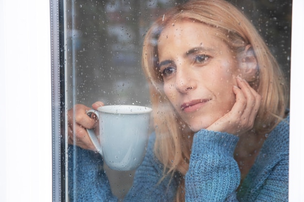 Reife Frau, die aus ihrem Fenster schaut, während es regnet und Tee trinkt