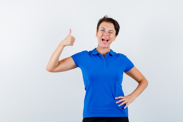 Reife frau, die daumen oben im blauen t-shirt zeigt und selbstbewusst, vorderansicht schaut.