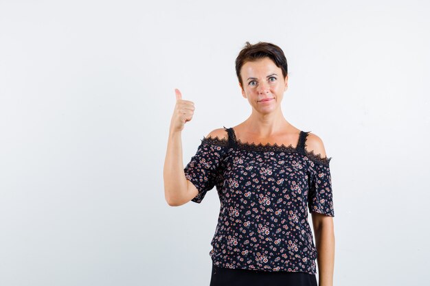 Reife Frau, die Daumen oben in der Blumenbluse, im schwarzen Rock zeigt und selbstbewusst aussieht. Vorderansicht.