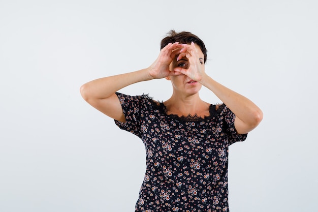 Reife Frau, die durch Hand als Fernglas in der Blumenbluse, im schwarzen Rock und in der fokussierten Vorderansicht schaut.