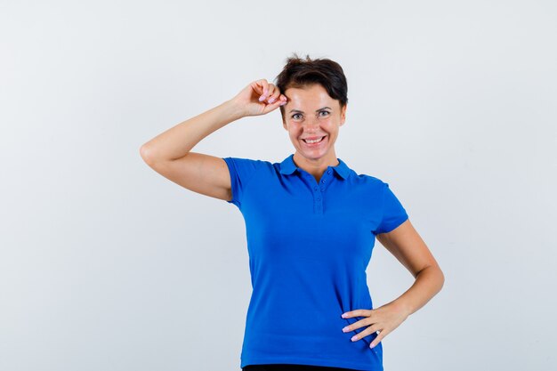 Reife Frau, die erhobene Hand auf Kopf im blauen T-Shirt hält und zuversichtlich schaut. Vorderansicht.