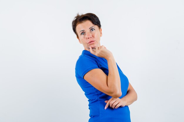 Reife Frau, die Finger am Kinn im blauen T-Shirt hält und verwirrt schaut. Vorderansicht.