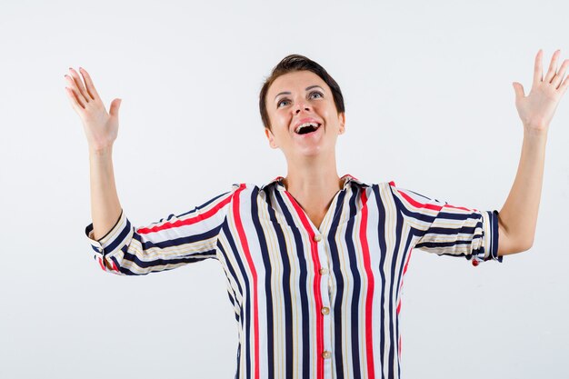 Reife Frau, die Hände als Freude im gestreiften Hemd erhebt und glücklich schaut. Vorderansicht.