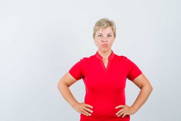 Reife Frau, die Hände auf Taille im roten T-Shirt hält und ernst schaut.