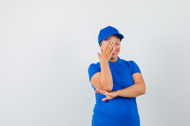 Reife Frau, die Hand auf einem Auge im blauen T-Shirt hält und freudig schaut