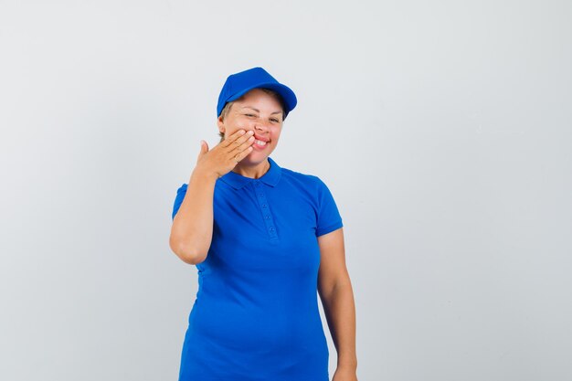 Reife Frau, die Hand auf Wange im blauen T-Shirt drückt und froh aussieht.