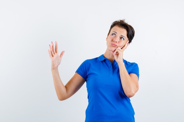 Reife Frau, die Hand hebt, während sie im blauen T-Shirt denkt und zweifelhafte Vorderansicht schaut.