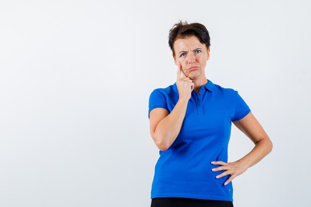 Reife Frau, die ihr Augenlid herunterzieht, während sie im blauen T-Shirt denkt und traurig, Vorderansicht schaut.