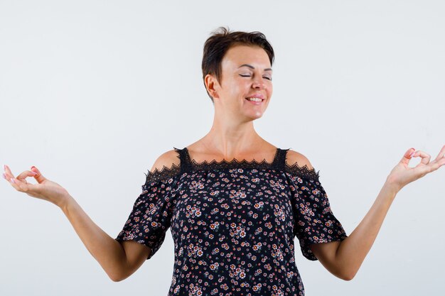 Reife frau, die in der meditierenden haltung in der blumenbluse, im schwarzen rock und in der entspannten vorderansicht steht.