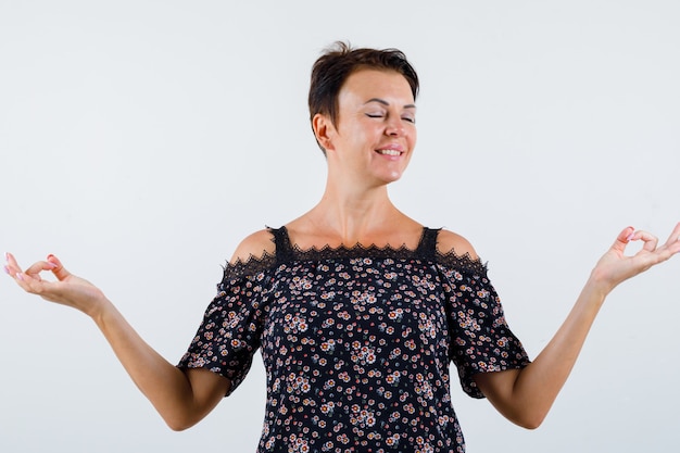 Reife Frau, die in der meditierenden Haltung in der Blumenbluse, im schwarzen Rock und in der entspannten Vorderansicht steht.