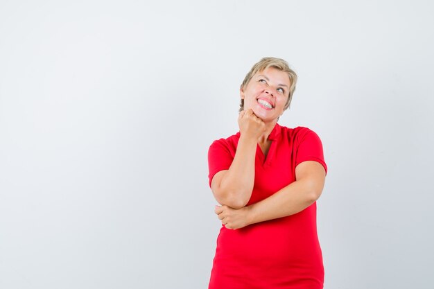 Reife Frau, die Kinn auf erhobene Faust im roten T-Shirt stützt und hoffnungsvoll aussieht.