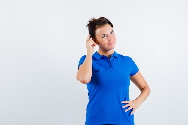 Reife Frau, die Kopf im blauen T-Shirt kratzt und verträumte Vorderansicht schaut.