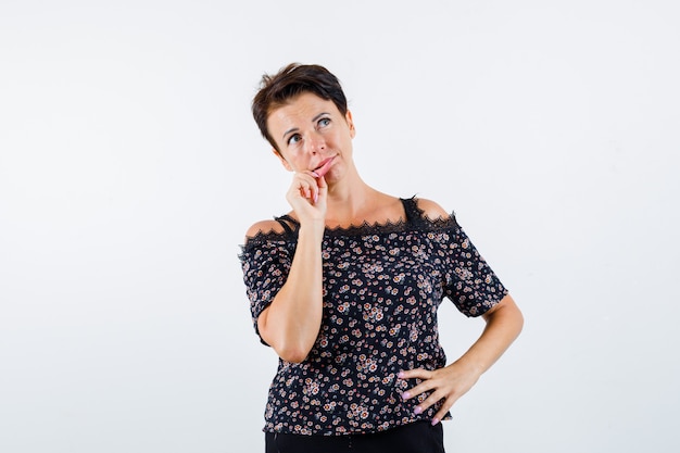 Reife Frau, die Lippen mit den Fingern zieht, Hand auf Taille in Blumenbluse, schwarzem Rock hält und nachdenklich aussieht. Vorderansicht.