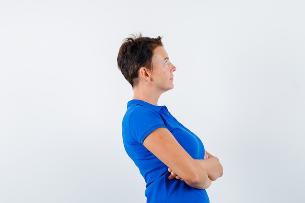 Reife Frau, die mit verschränkten Armen im blauen T-Shirt steht und nachdenklich schaut.