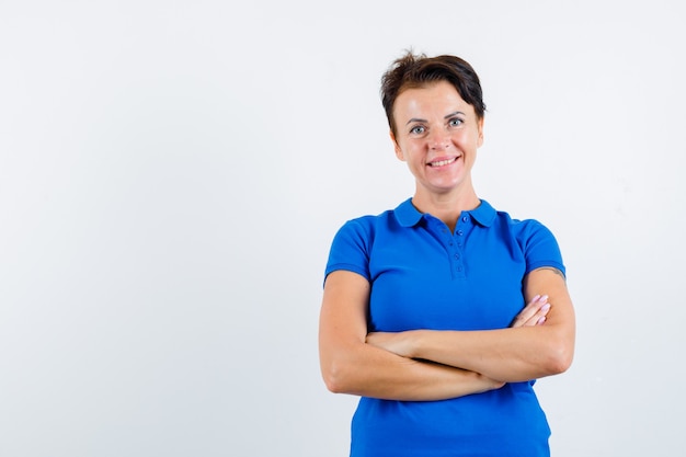 Reife Frau, die mit verschränkten Armen im blauen T-Shirt steht und selbstbewusst, Vorderansicht schaut.