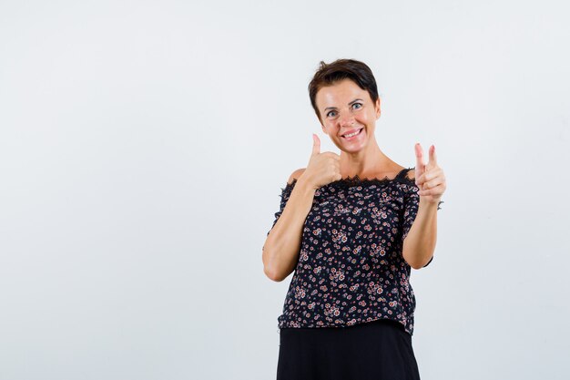 Reife Frau, die mit Zeigefinger auf Kamera zeigt, Daumen oben in Blumenbluse, schwarzem Rock und fröhlich aussehend, Vorderansicht zeigend.