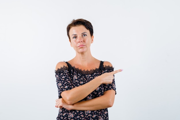 Reife Frau, die mit Zeigefinger in Blumenbluse und schwarzem Rock nach rechts zeigt und ernsthafte Vorderansicht sieht.