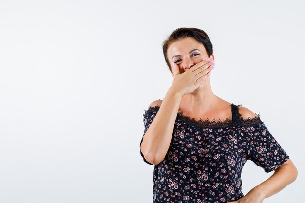 Reife Frau, die Mund mit Hand bedeckt, Hand auf Taille in der Blumenbluse, im schwarzen Rock hält und glücklich schaut. Vorderansicht.