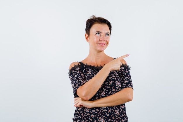 Reife Frau, die rechts mit dem Zeigefinger in der Blumenbluse, im schwarzen Rock zeigt und lustig schaut, Vorderansicht.