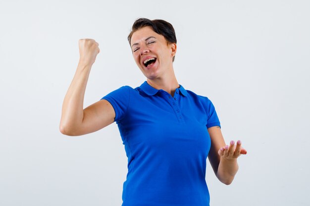 Reife Frau, die Siegergeste im blauen T-Shirt zeigt und glücklich schaut. Vorderansicht.