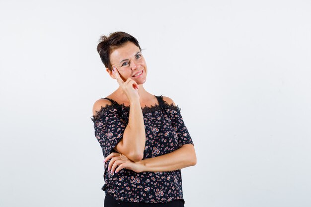 Reife Frau, die Wange auf Handfläche lehnt, an etwas in der Blumenbluse, im schwarzen Rock denkt und glücklich aussieht. Vorderansicht.