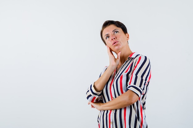 Reife Frau, die Wange auf Handfläche lehnt, eine Hand auf Ellbogen in gestreifter Bluse hält und nachdenklich aussieht. Vorderansicht.