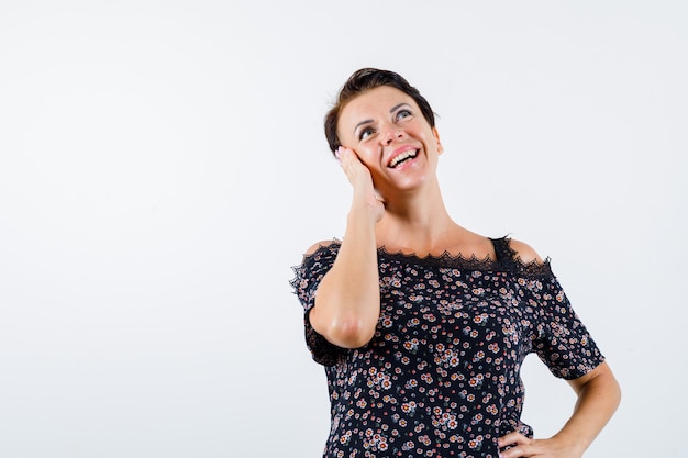 Reife Frau, die Wange auf Handfläche lehnt, Hand auf Taille hält, in Blumenbluse, schwarzem Rock nach oben schauend und fröhlich, Vorderansicht schauend.