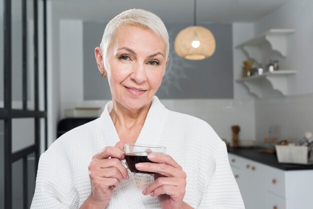 Reife Frau im Bademantel, der in der Küche beim Halten der Kaffeetasse aufwirft