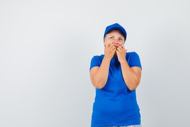 Reife Frau im blauen T-Shirt beißt Fäuste emotional und sieht ängstlich aus.