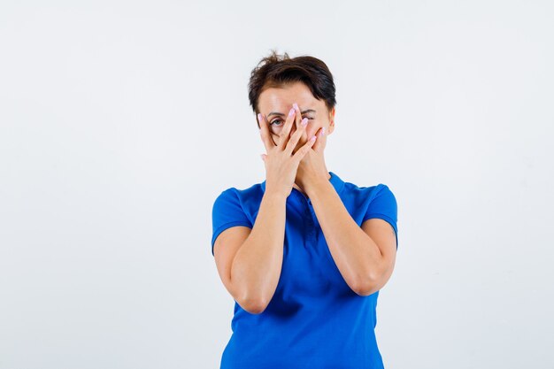 Reife Frau im blauen T-Shirt, das durch Finger schaut und neugierige Vorderansicht schaut.