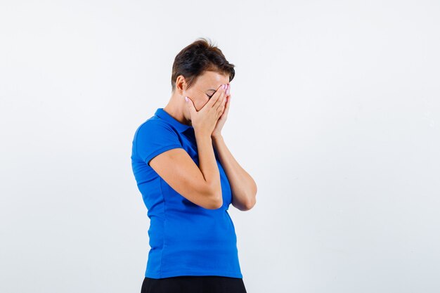 Reife Frau im blauen T-Shirt, das Gesicht mit Händen bedeckt und deprimiert, Vorderansicht schaut.